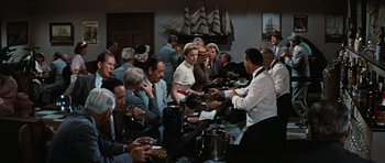 Movie still from “Peyton Place” (1957), directed by Mark Robson – A group of people standing around a table with plates of food on top of it; Wide shot, High angle