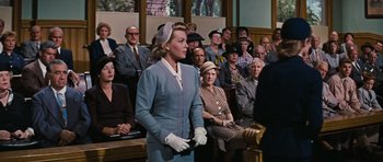 Movie still from “Peyton Place” (1957), directed by Mark Robson – A woman standing in front of a crowd of people in a courtroom; Medium shot, Over the shoulder angle
