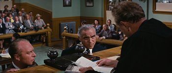 Movie still from “Peyton Place” (1957), directed by Mark Robson – An older man in a suit and tie sitting at a table in a courtroom; Medium shot, Over the shoulder angle