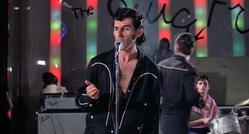 Movie still from “Phantom of the Paradise” (1974), directed by Brian De Palma – A man in black jacket holding a microphone in front of a crowd; Medium shot, Low angle