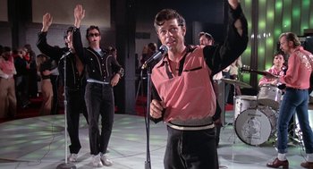 Movie still from “Phantom of the Paradise” (1974), directed by Brian De Palma – A man holding a microphone in front of a crowd; Medium shot, Low angle