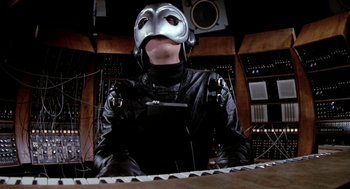 Movie still from “Phantom of the Paradise” (1974), directed by Brian De Palma – A person wearing a mask and a leather jacket sitting in front of a keyboard; Close Up shot, Low angle