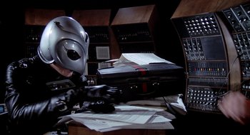 Movie still from “Phantom of the Paradise” (1974), directed by Brian De Palma – A person in a mask sitting at a desk; Medium shot, High angle