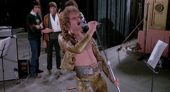 Movie still from “Phantom of the Paradise” (1974), directed by Brian De Palma – A man in a gold suit is singing into a microphone; Medium shot, Low angle