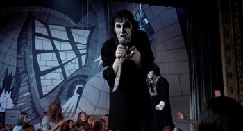 Movie still from “Phantom of the Paradise” (1974), directed by Brian De Palma – A man with a knife in his mouth standing in front of a crowd of onlookers; Medium shot, Low angle