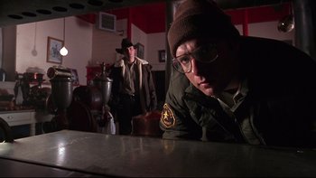 Movie still from “Phantoms” (1998), directed by Joe Chappelle – A man in a hat and glasses is sitting at a table with another man in a hat and glasses; Medium shot, Low angle