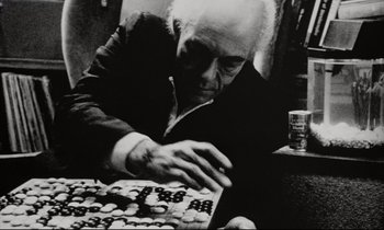 Movie still from “Pi” (1998), directed by Darren Aronofsky – An older man playing a game of checkers in a room; Medium shot, Low angle