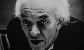 Movie still from “Pi” (1998), directed by Darren Aronofsky – An old man with a white beard and a black shirt; Close Up shot, Low angle