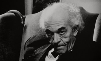 Movie still from “Pi” (1998), directed by Darren Aronofsky – An older man sitting in a chair looking at the camera; Close Up shot, High angle