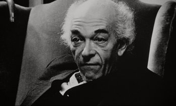 Movie still from “Pi” (1998), directed by Darren Aronofsky – An older man sitting on a couch smoking a cigarette; Close Up shot, Low angle