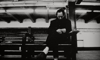 Movie still from “Pi” (1998), directed by Darren Aronofsky – A man sitting on top of a wooden park bench; Medium shot, Low angle