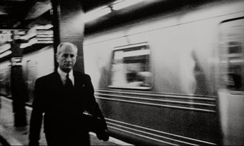Movie still from “Pi” (1998), directed by Darren Aronofsky – A black and white photo of a man in a suit and tie standing in front of a train; Medium shot, Low angle