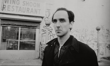 Movie still from “Pi” (1998), directed by Darren Aronofsky – Black and white photograph of a man standing in front of a building; Close Up shot, Low angle
