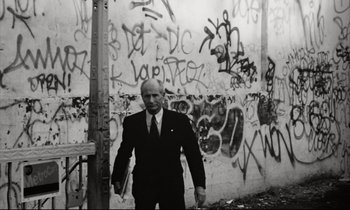 Movie still from “Pi” (1998), directed by Darren Aronofsky – A black and white photo of a man in a suit and tie in front of a wall covered in graffiti; Medium shot, Low angle
