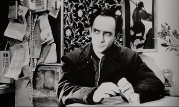 Movie still from “Pi” (1998), directed by Darren Aronofsky – A black and white photo of a man sitting at a table; Close Up shot, High angle