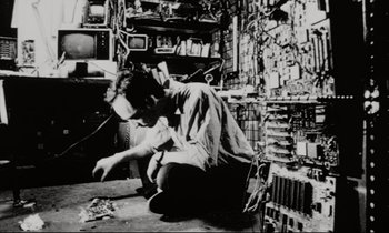 Movie still from “Pi” (1998), directed by Darren Aronofsky – A man sitting on the ground in a room filled with lots of stuff; Medium shot, Low angle
