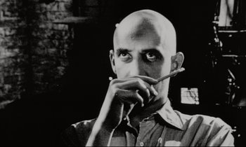 A man with a shaved head holding a pen in his mouth