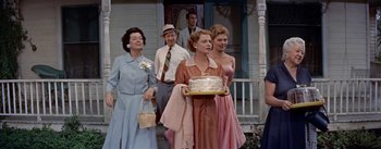 Movie still from “Picnic” (1955), directed by Joshua Logan – A group of people standing on a porch holding a cake; Medium shot, Over the shoulder angle