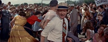 Movie still from “Picnic” (1955), directed by Joshua Logan – A man in a hat and a tie is standing in front of a group of people; Medium shot, Over the shoulder angle