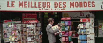 Movie still from “Pierrot le Fou” (1965), directed by Jean-Luc Godard – A man is standing in front of a store holding a book; Medium shot, Low angle