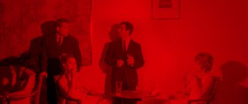 Movie still from “Pierrot le Fou” (1965), directed by Jean-Luc Godard – A man in a suit standing next to a table with a glass of water; Medium shot, High angle