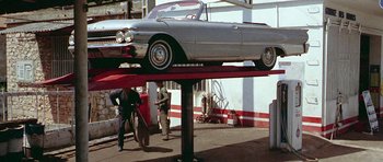 Movie still from “Pierrot le Fou” (1965), directed by Jean-Luc Godard – Two men working on a car on a car lift; Wide shot, Low angle