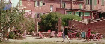 Movie still from “Pierrot le Fou” (1965), directed by Jean-Luc Godard – A woman walking in front of a pink building with lawn chairs; Extreme Wide shot, High angle