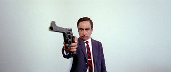 Movie still from “Pierrot le Fou” (1965), directed by Jean-Luc Godard – A man in a suit and tie holding a gun pointed at the camera; Medium shot, Low angle
