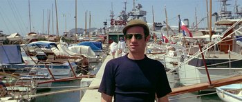 Movie still from “Pierrot le Fou” (1965), directed by Jean-Luc Godard – A man wearing a hat standing in front of boats; Medium shot, Low angle