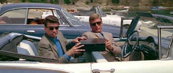 Movie still from “Pierrot le Fou” (1965), directed by Jean-Luc Godard – Two men sitting in the back seat of a car; Medium shot, Low angle