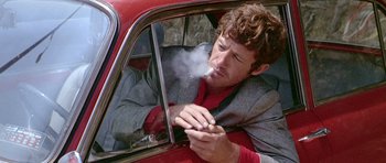 Movie still from “Pierrot le Fou” (1965), directed by Jean-Luc Godard – A man smoking a cigarette while sitting in the passenger seat of a car; Close Up shot, Low angle