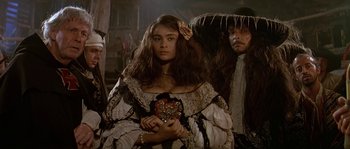Movie still from “Pirates” (1986), directed by Roman Polanski – A woman in a dress holding a heart in front of a group of people; Close Up shot, Over the shoulder angle