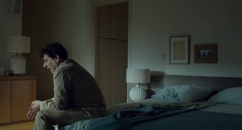 Movie still from “Pity” (2018), directed by Babis Makridis – A man sitting on the edge of a bed in a bedroom; Medium shot, Low angle