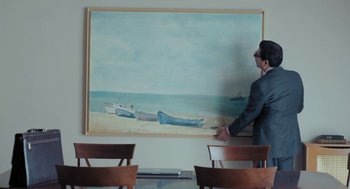 Movie still from “Pity” (2018), directed by Babis Makridis – A man standing in front of a large painting of a boat; Medium shot, High angle