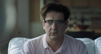 Movie still from “Pity” (2018), directed by Babis Makridis – A person wearing glasses and a pink shirt; Close Up shot, Over the shoulder angle