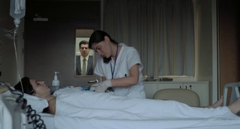 Movie still from “Pity” (2018), directed by Babis Makridis – A woman in a hospital gown is working on a patient; Medium shot, Over the shoulder angle