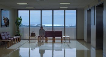 Movie still from “Pity” (2018), directed by Babis Makridis – A woman sitting on a bench in front of a large window; Extreme Wide shot, Low angle