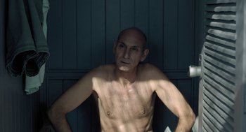 Movie still from “Pity” (2018), directed by Babis Makridis – A shirtless man sitting in a bathroom stall; Medium shot, High angle