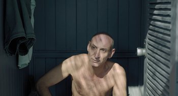 Movie still from “Pity” (2018), directed by Babis Makridis – A shirtless man sitting in front of a wooden wall; Medium shot, Low angle