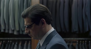 Movie still from “Pity” (2018), directed by Babis Makridis – A person wearing a suit and glasses; Close Up shot, Over the shoulder angle