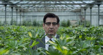 Movie still from “Pity” (2018), directed by Babis Makridis – A man wearing glasses and a suit is surrounded by plants; Medium shot, Over the shoulder angle