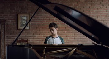 Movie still from “Pity” (2018), directed by Babis Makridis – A young man sitting in front of an open piano; Medium shot, High angle