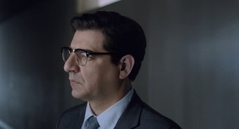 Movie still from “Pity” (2018), directed by Babis Makridis – A person wearing glasses and a suit; Close Up shot, Over the shoulder angle