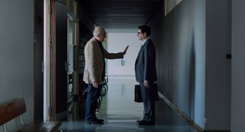 Movie still from “Pity” (2018), directed by Babis Makridis – Two men in a hallway one of which is holding his hand up to the other; Medium shot, Over the shoulder angle
