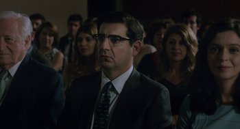 Movie still from “Pity” (2018), directed by Babis Makridis – A man in a suit and tie sitting in front of a group of people; Close Up shot, Over the shoulder angle