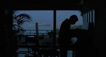 Movie still from “Pity” (2018), directed by Babis Makridis – A man standing at a table in front of an ocean view; Wide shot, Low angle
