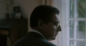Movie still from “Pity” (2018), directed by Babis Makridis – A person wearing glasses and a suit; Close Up shot, Over the shoulder angle