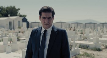 Movie still from “Pity” (2018), directed by Babis Makridis – A man wearing a suit and tie standing in front of a cemetery; Medium shot, Low angle