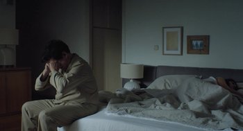 Movie still from “Pity” (2018), directed by Babis Makridis – A man sitting on top of a bed in a bedroom; Medium shot, High angle