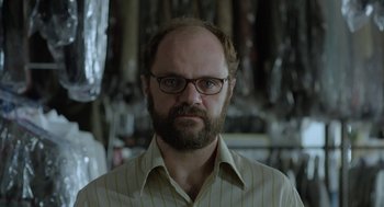 Movie still from “Pity” (2018), directed by Babis Makridis – A man with a goatee wearing glasses and a striped shirt; Close Up shot, Low angle